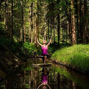 Foto mit besonderen Lichteffekten zeigt Eva beim Duchführen einer Fitnessübung im Wald, im Wasser spiegelnd.